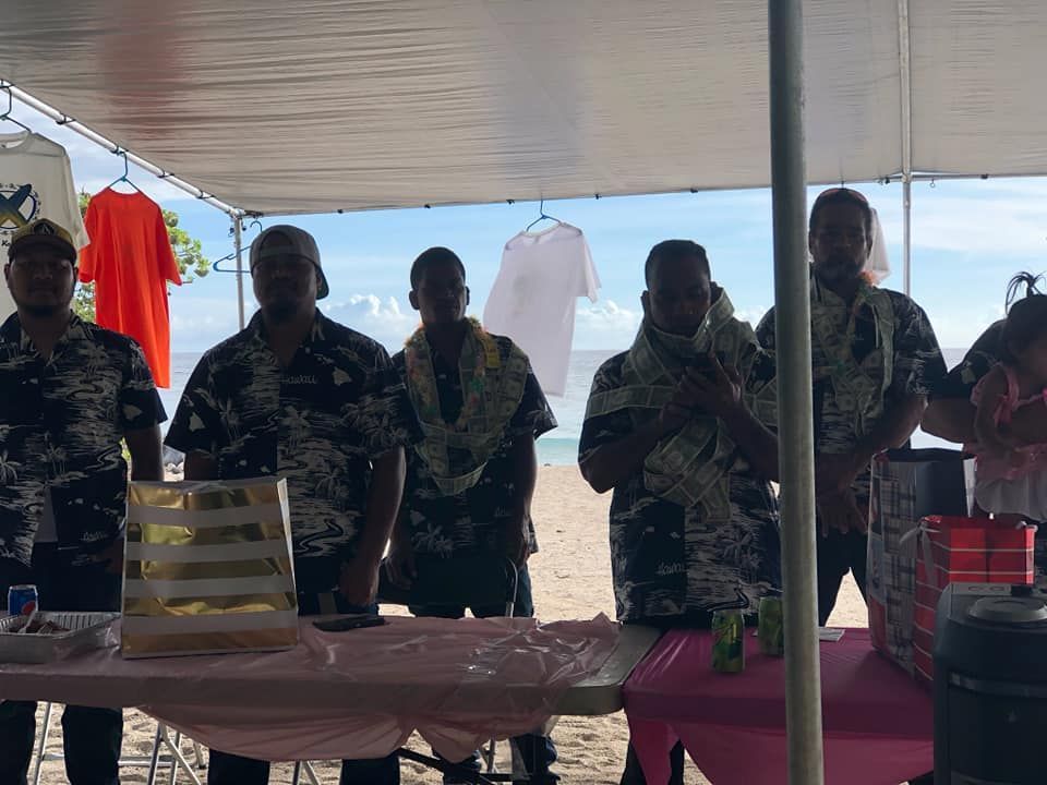 Men at a beach booth, wearing leis and floral shirts. Pink tablecloth. Two shirts hanging behind them.