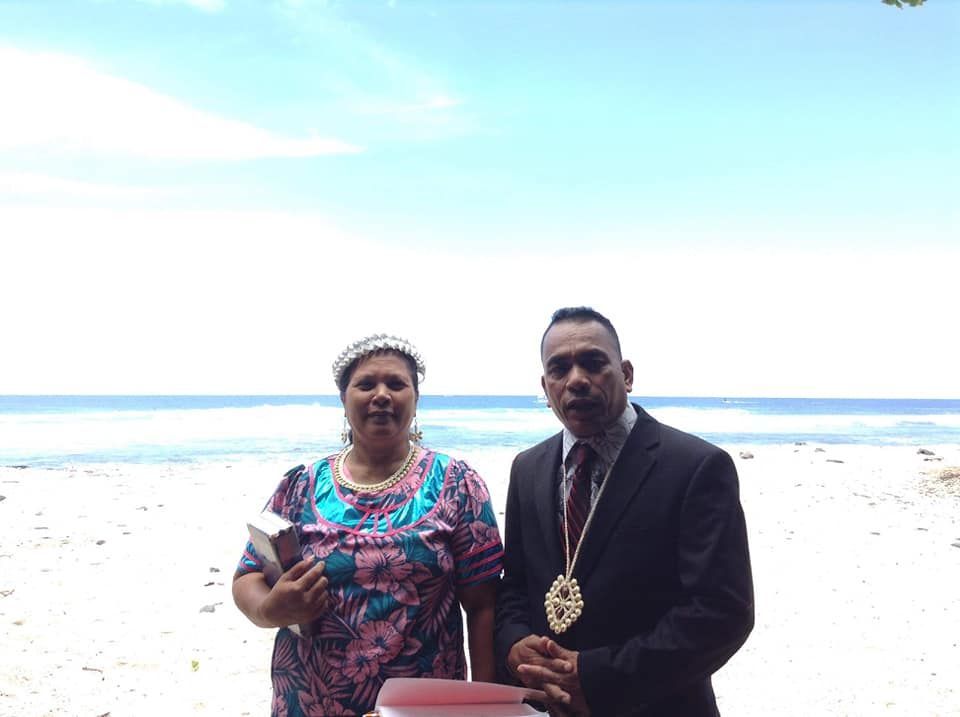 Two people stand on a beach. Woman holds book, wears floral dress and crown. Man in suit with gold chain.