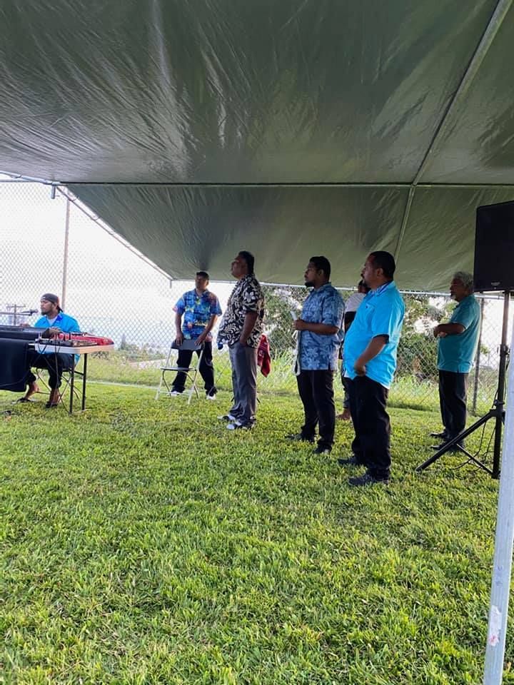 A group of men under a canopy on grass, some listening to a speaker at a table.