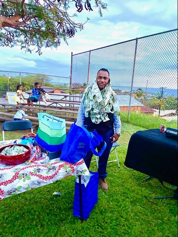 Person standing on grass, smiling, wearing a floral scarf, carrying blue bags, near a table, fence, and seating area.