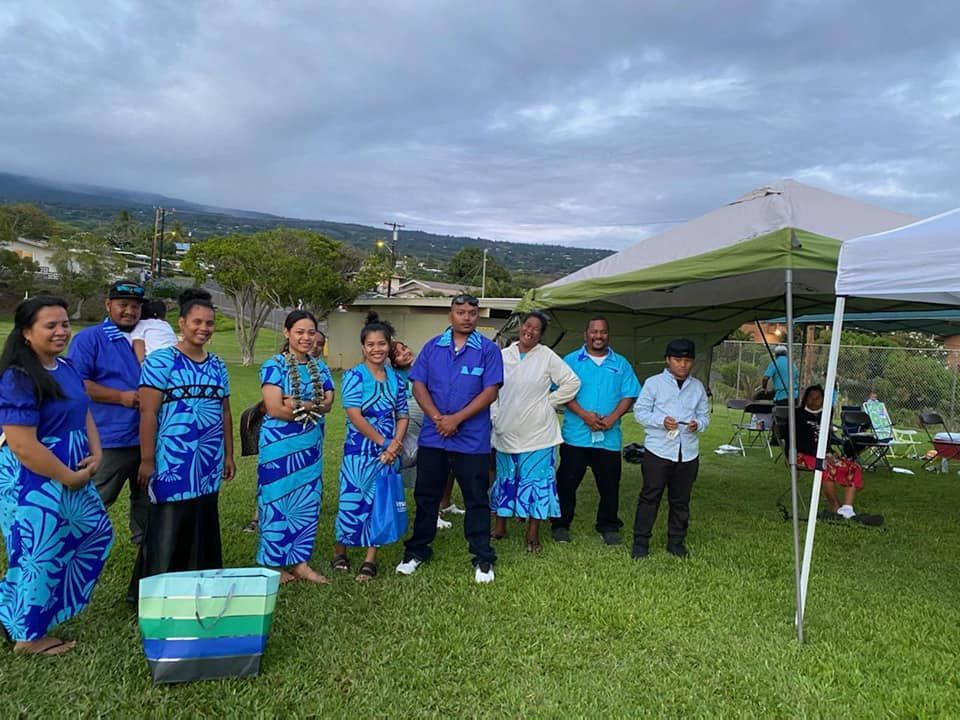 Group of people in blue attire stand in front of a tent on a grassy area with a mountain in the background.