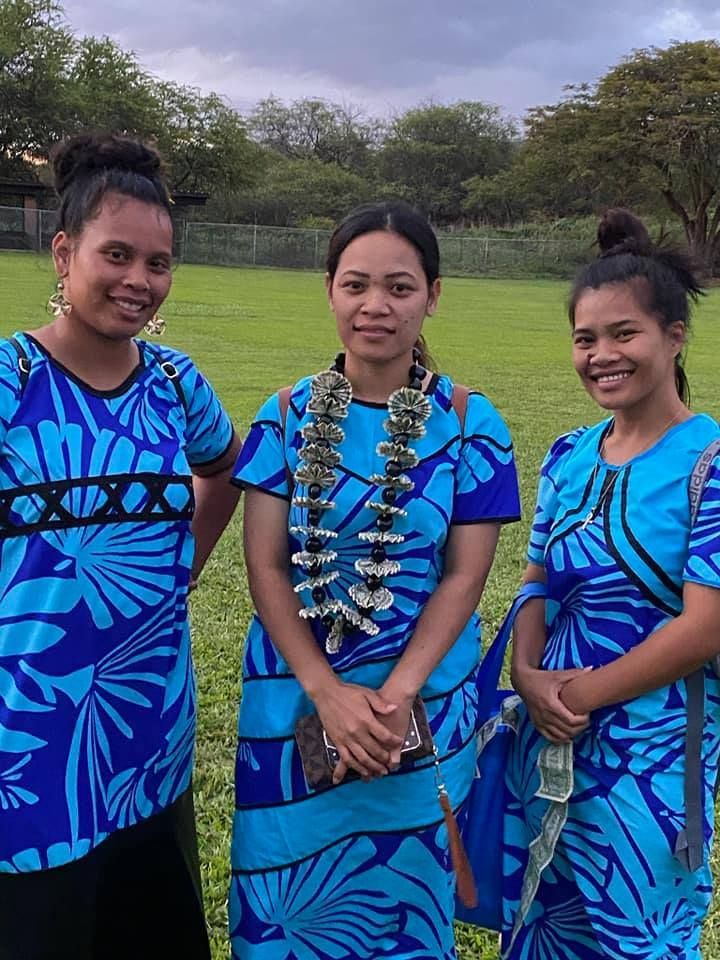 Three women in blue patterned dresses stand in a grassy area, smiling. One wears a lei.
