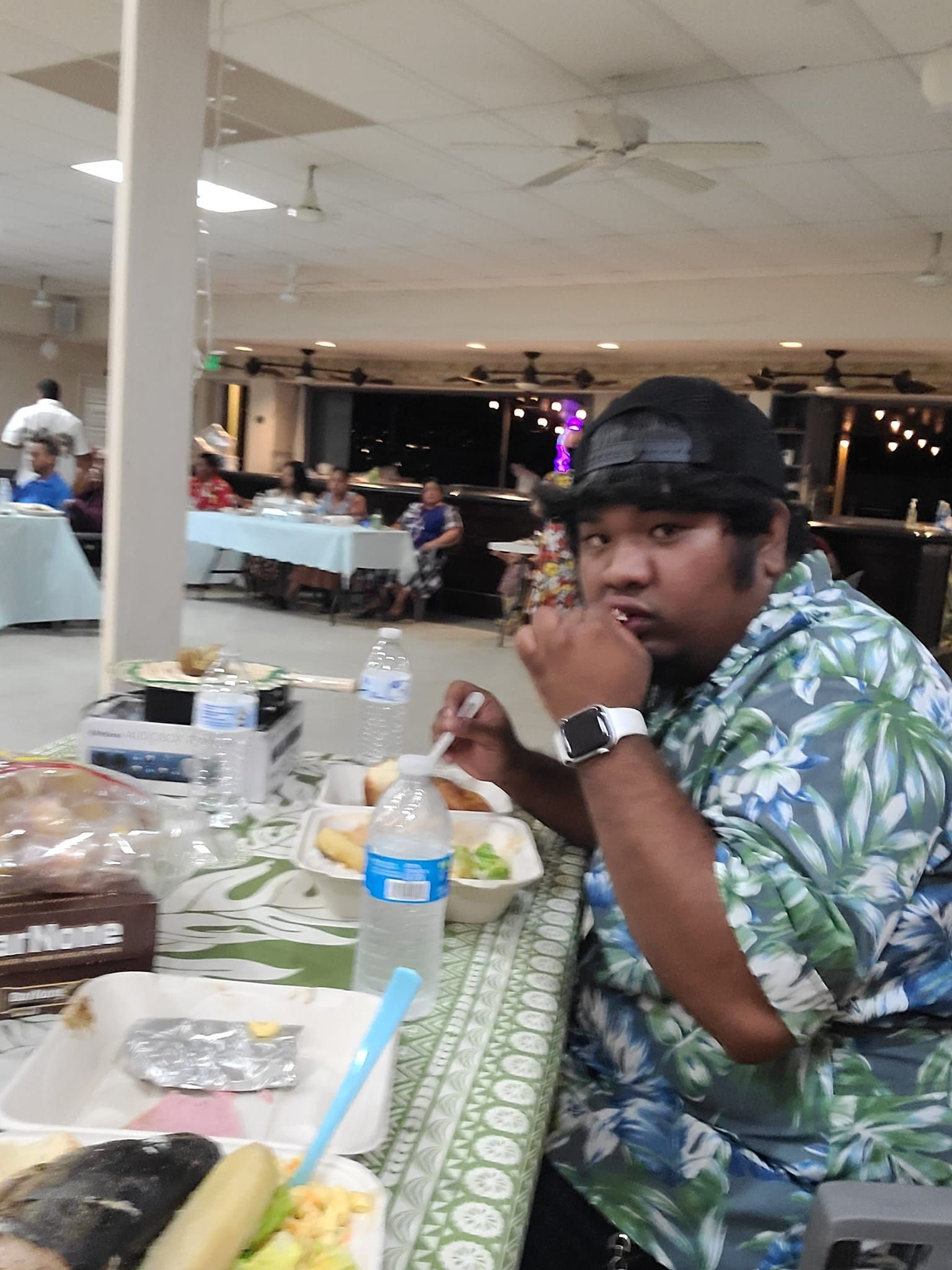 Man in floral shirt eats at a table in a hall, looking at camera.