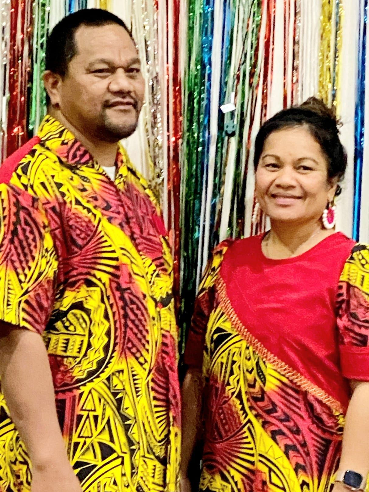 Couple in matching yellow and red patterned shirts, smiling, against colorful streamers.
