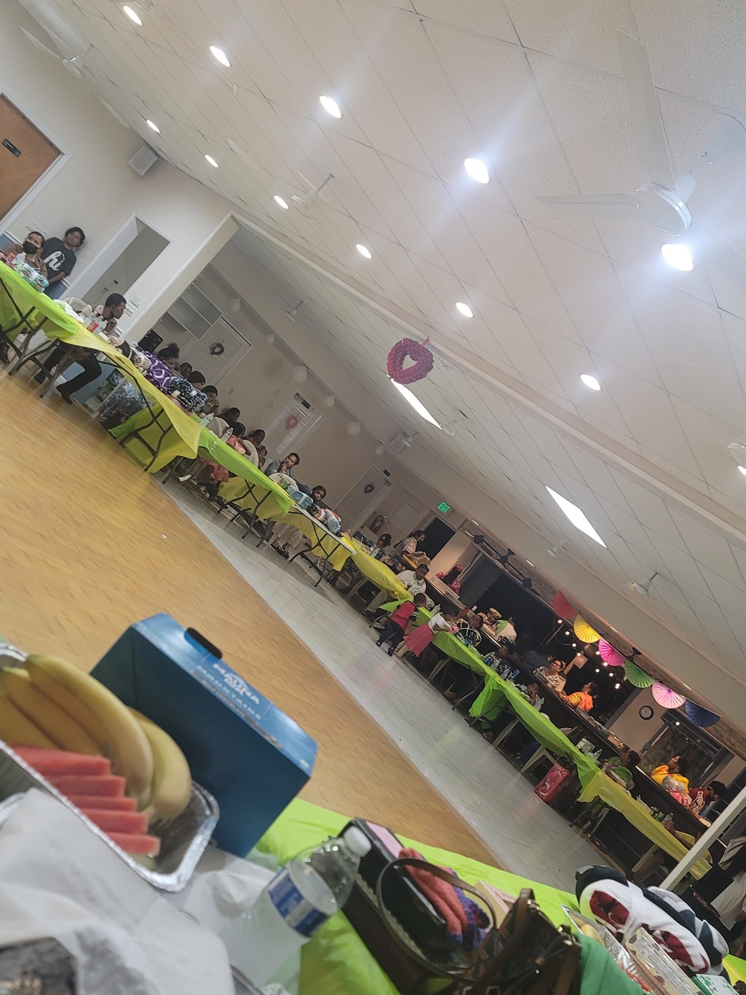 People at long tables in a brightly lit hall, with fruit and food in the foreground.