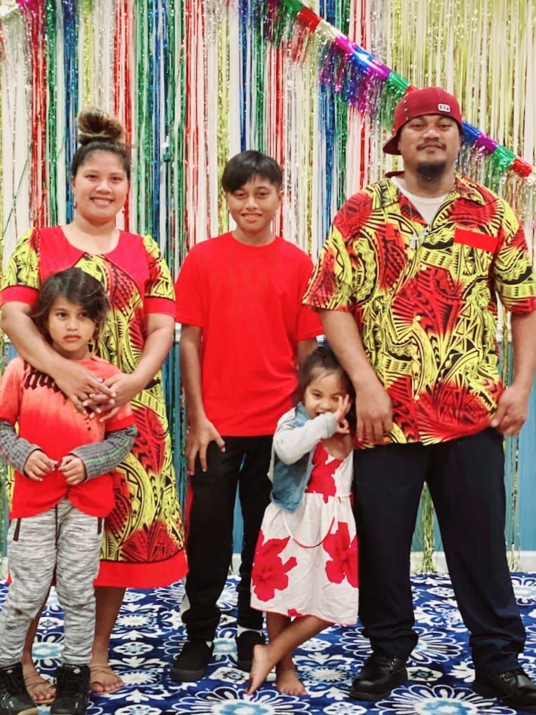 Family of five poses in front of colorful streamers. Woman in red dress, two children, teenage boy, and man in red shirt.