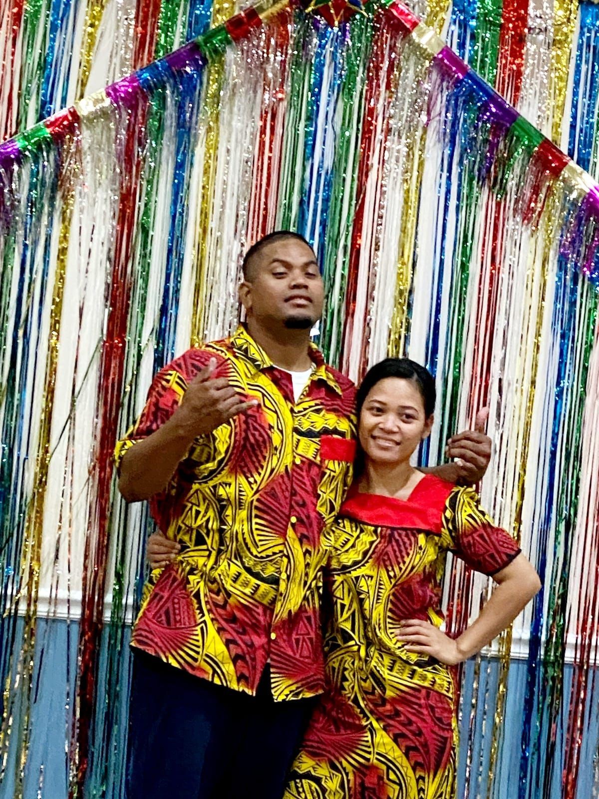 Man and woman in matching red and yellow patterned shirts posing in front of colorful streamers.