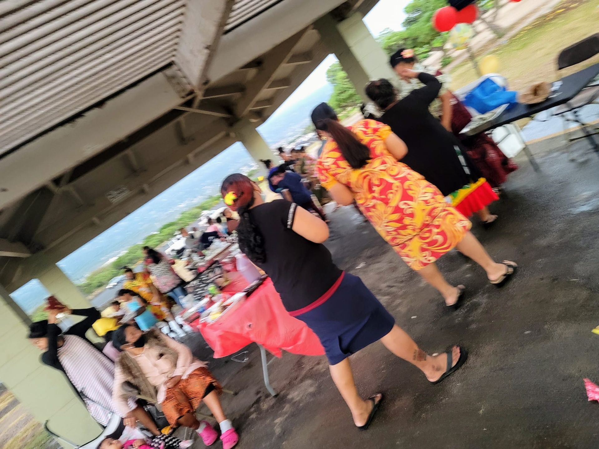 People at an outdoor gathering under a shelter. Some are dancing. Red, orange, and black clothing.