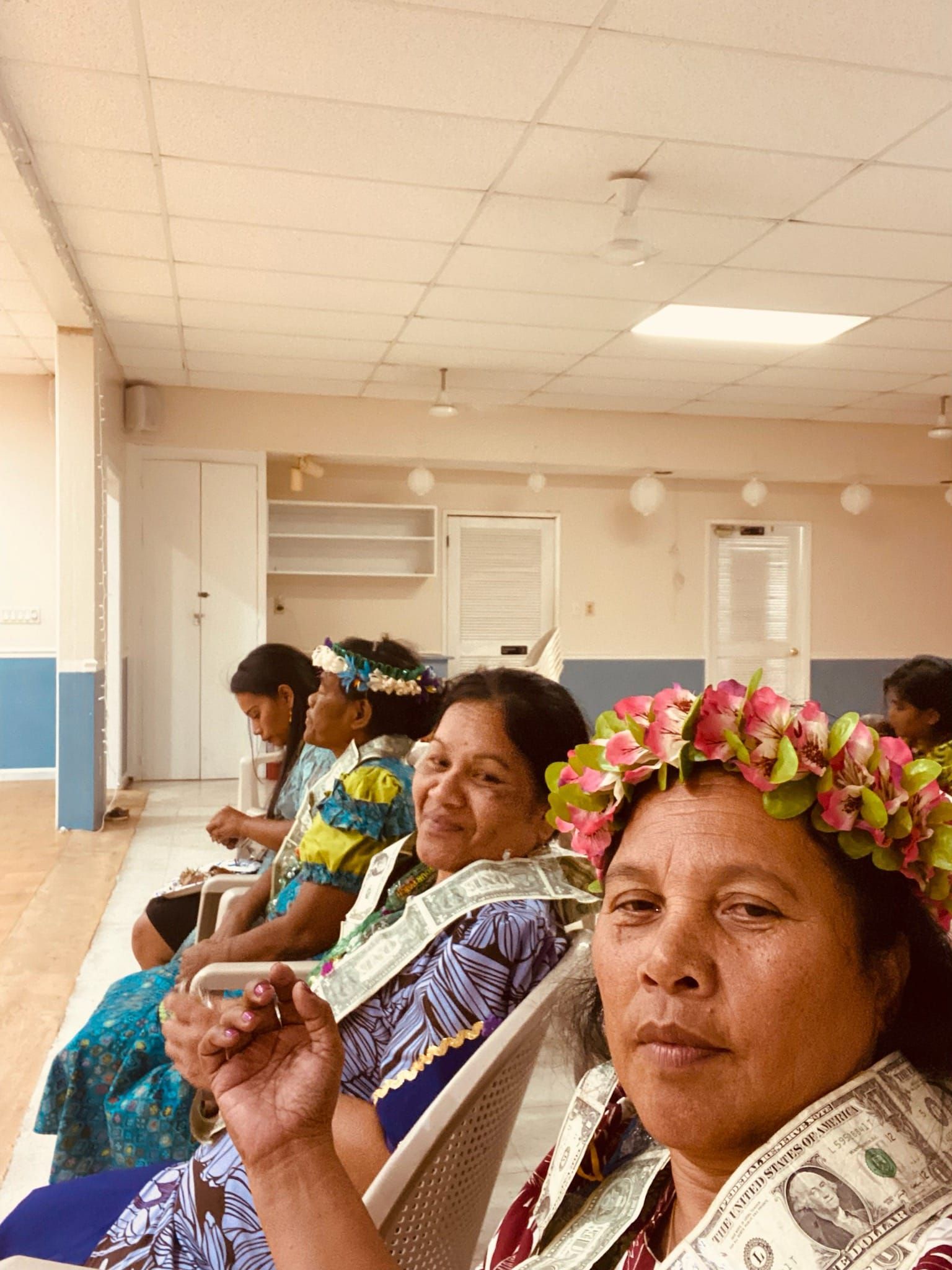 Women in a room, some wearing flower crowns and lei with money.