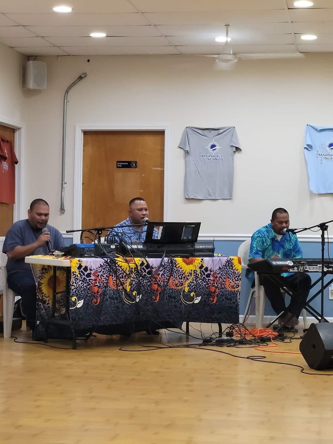 Three men performing with keyboards, microphones in a room. One table has a colorful cloth.