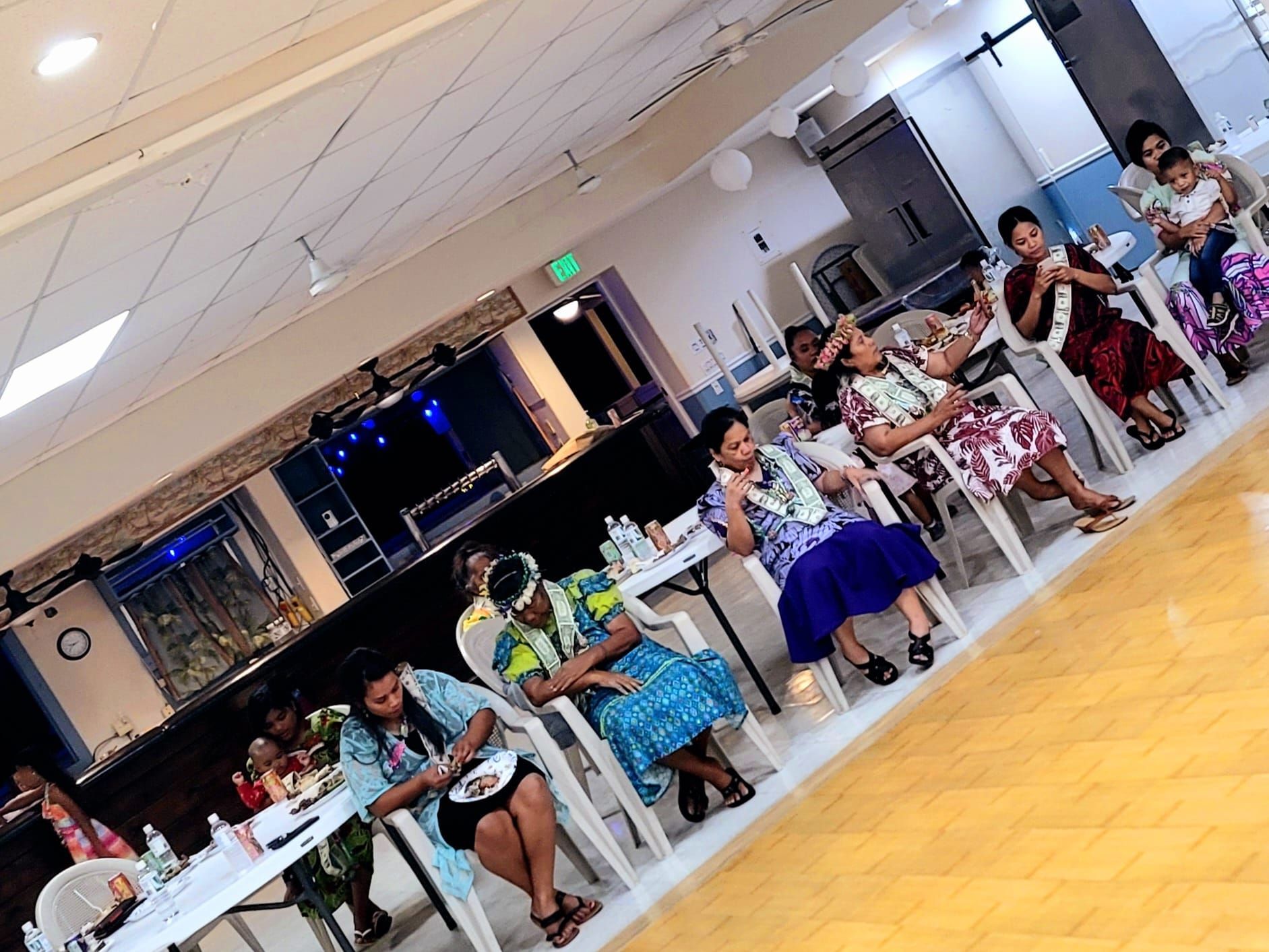 People seated in chairs at a gathering, wearing leis and patterned clothing. Indoors, tables with food.