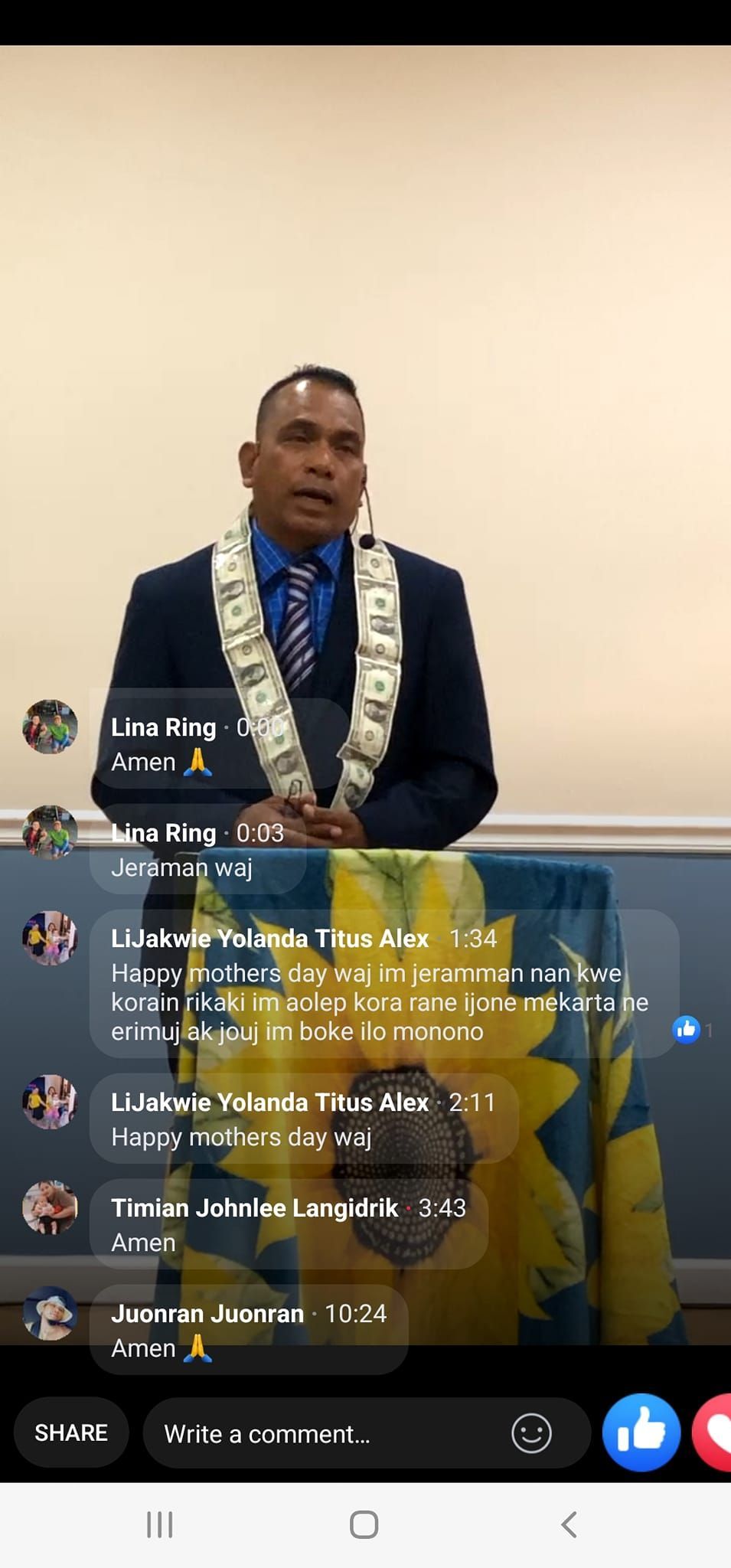 A man speaking at a podium with a sunflower design, live streaming. He wears a blue suit and a lei.