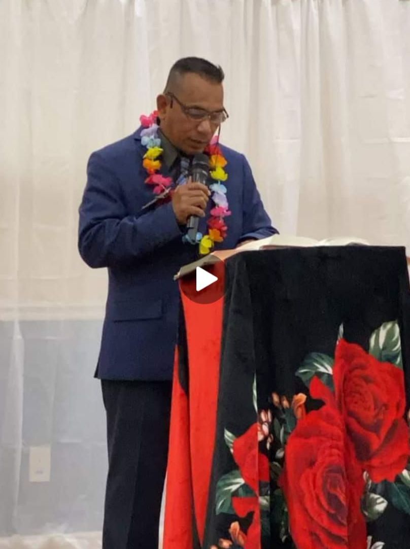 Man in blue suit and lei reads from a podium with a floral cover.
