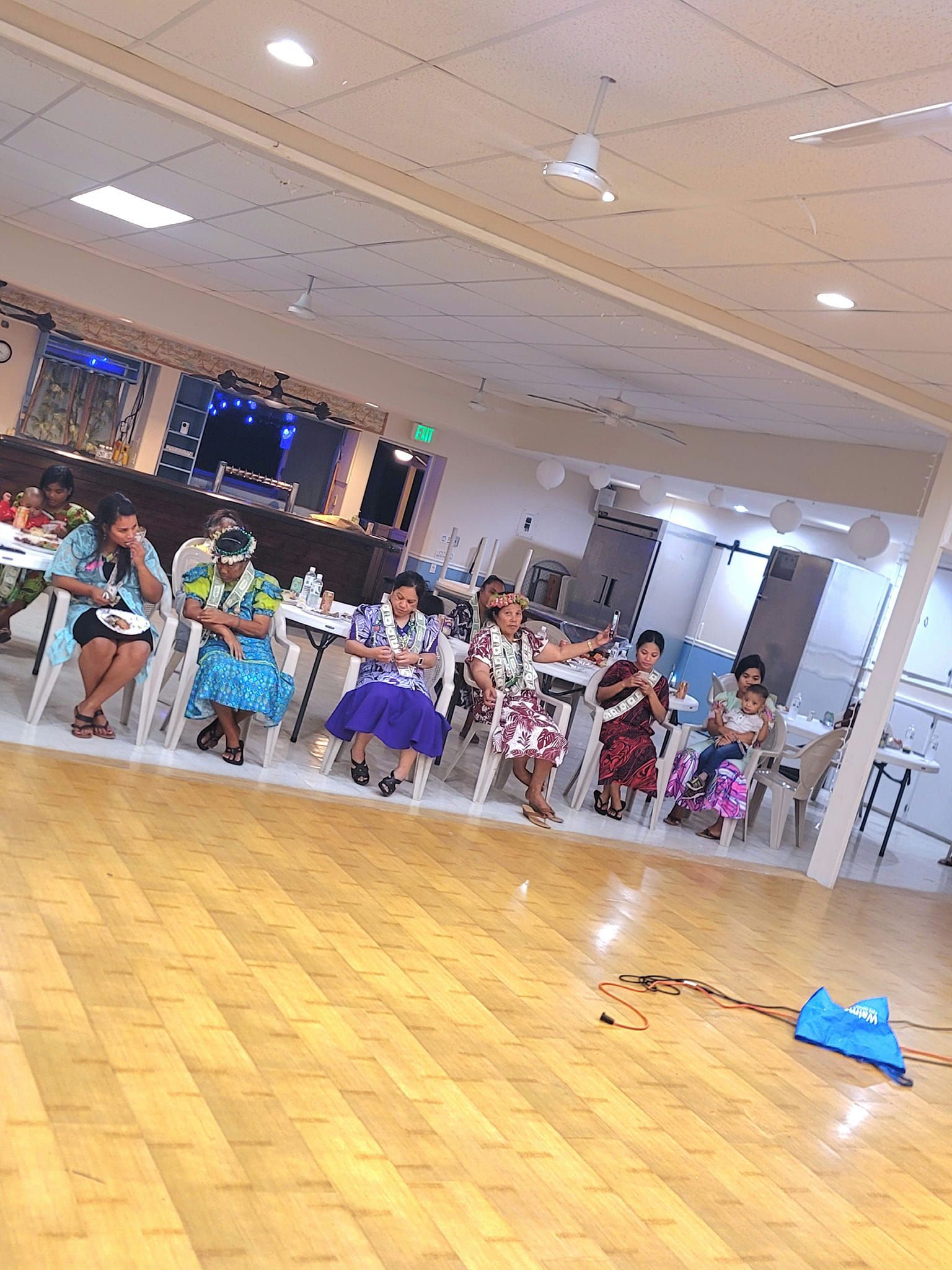 Women in colorful dresses and leis sit at a table, some with hands raised, in a well-lit room.