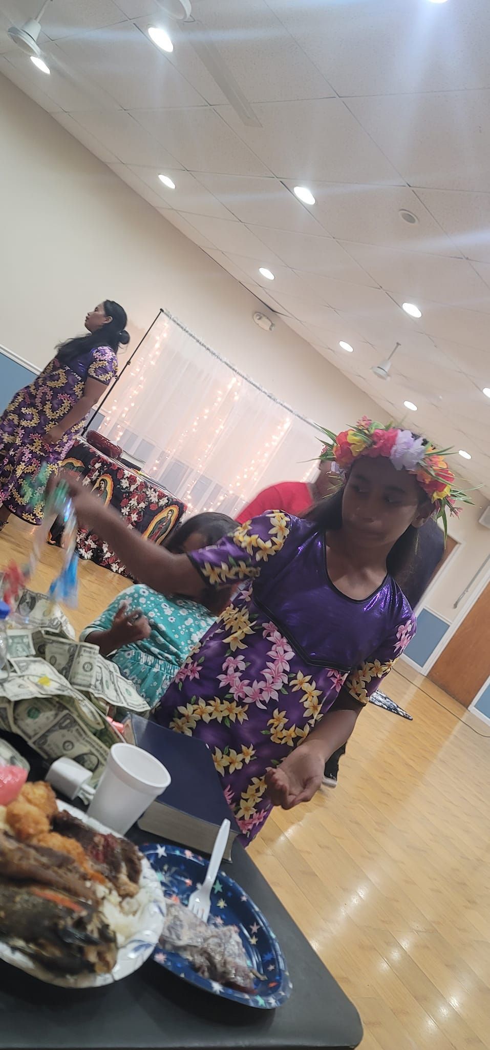 Woman with flower crown, floral dress, and ribbon at event, surrounded by food and people.