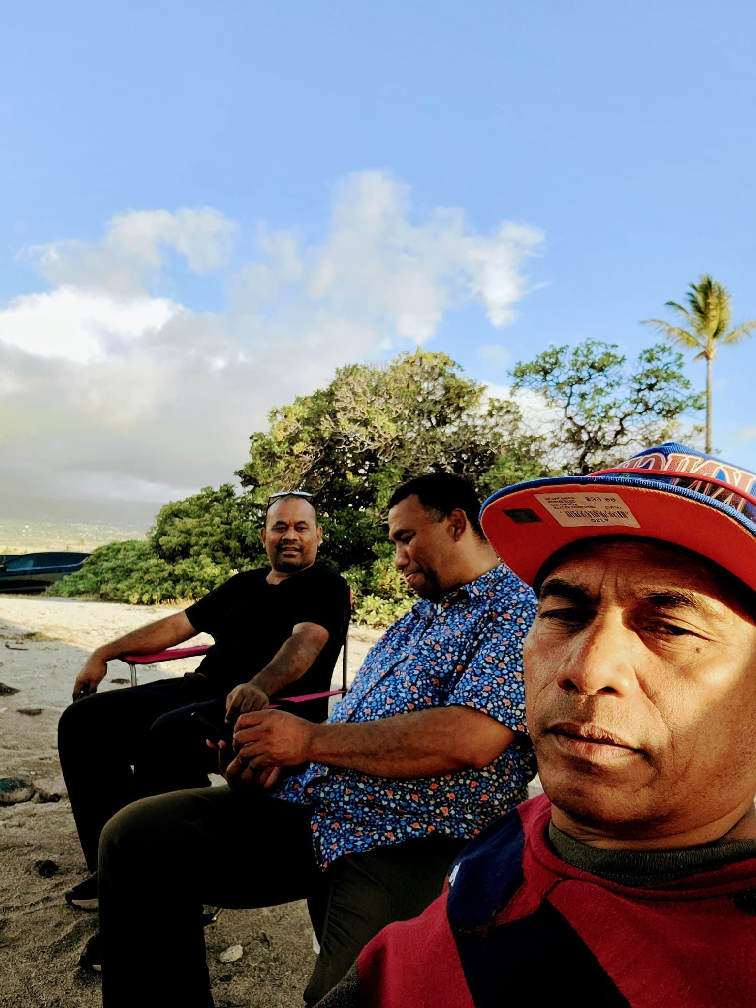 Three men sitting outside on a cloudy day. One wearing a cap, two in shirts, one with dark hair.