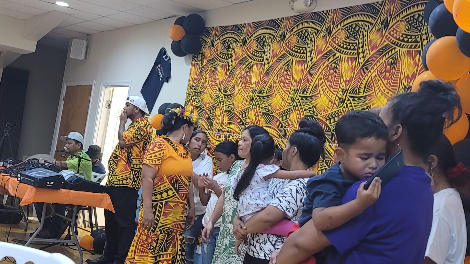 People in a brightly decorated room; some in traditional clothing, others holding children. Balloons decorate the background.