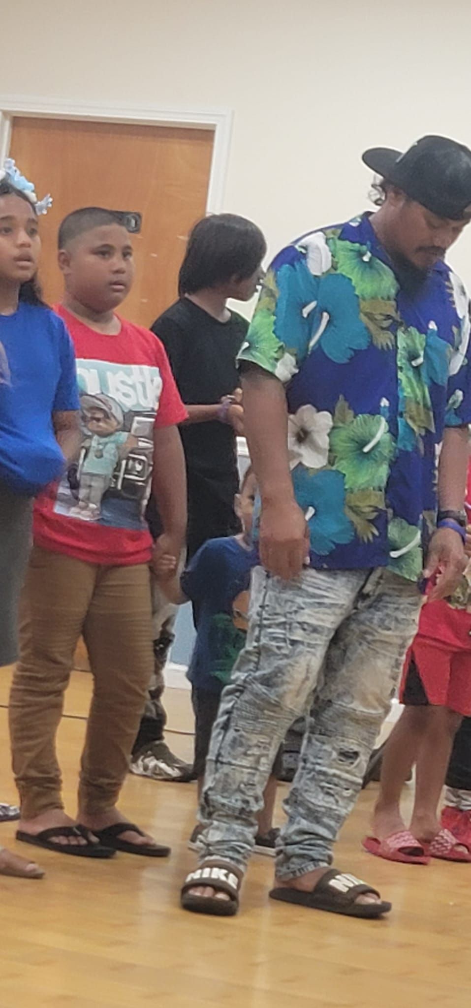 A group of people, including children and a man in patterned shirt and jeans, stand in a room.