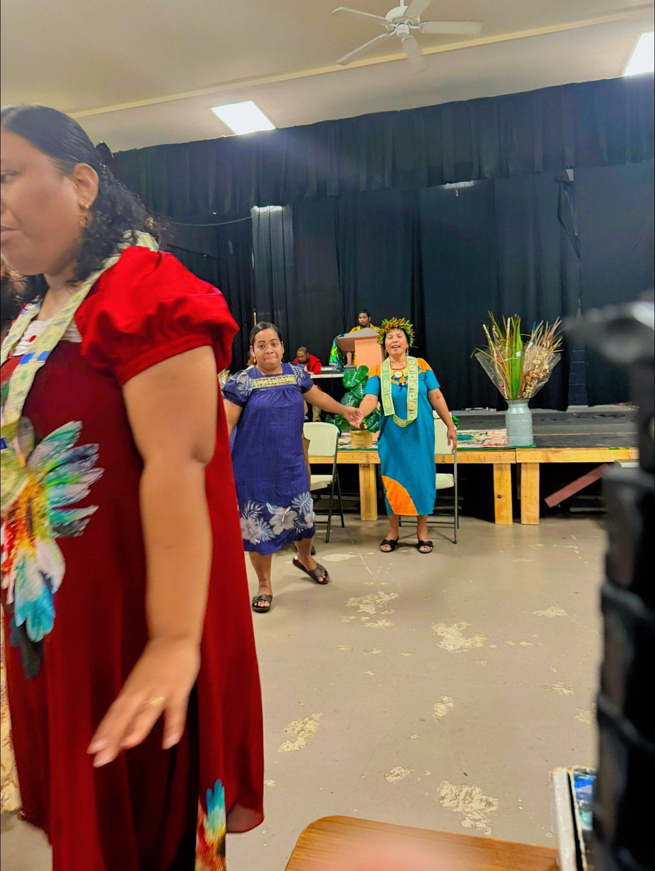 Three women in colorful dresses dancing onstage, decorated with flower leis.