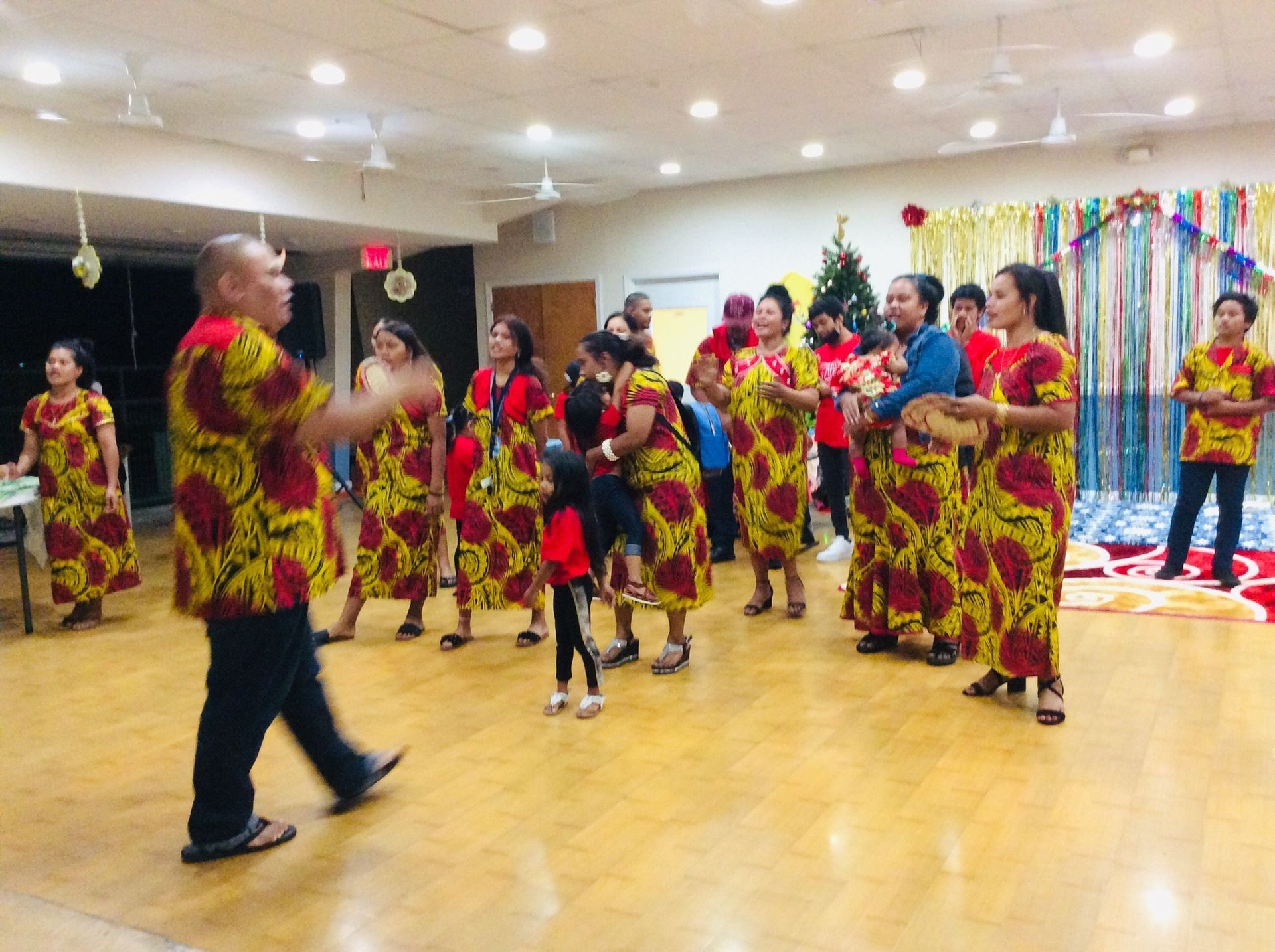 People in matching yellow-red dresses dance in a brightly lit room with a Christmas tree.