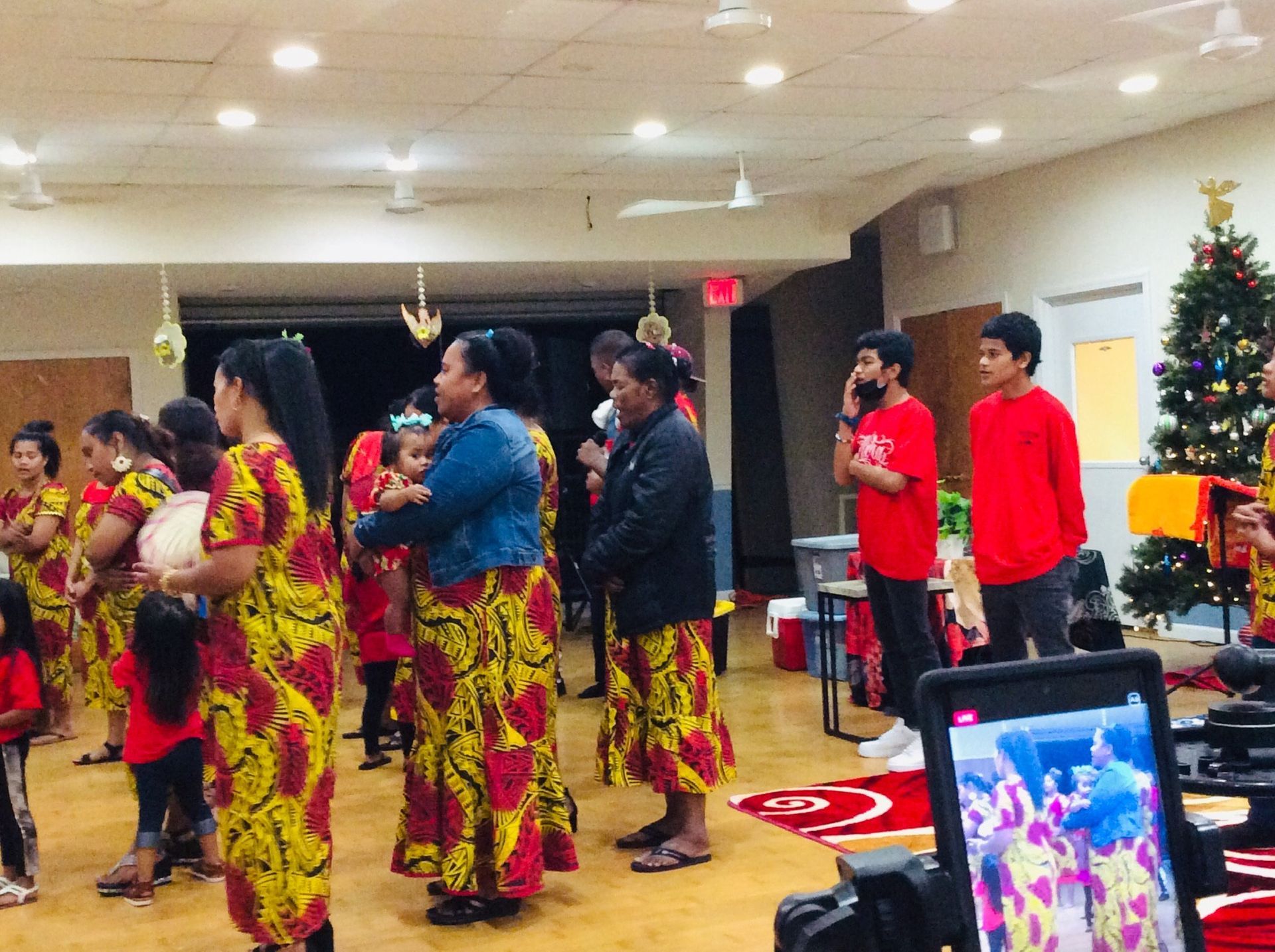 People in colorful clothing stand indoors, some with a Christmas tree visible.