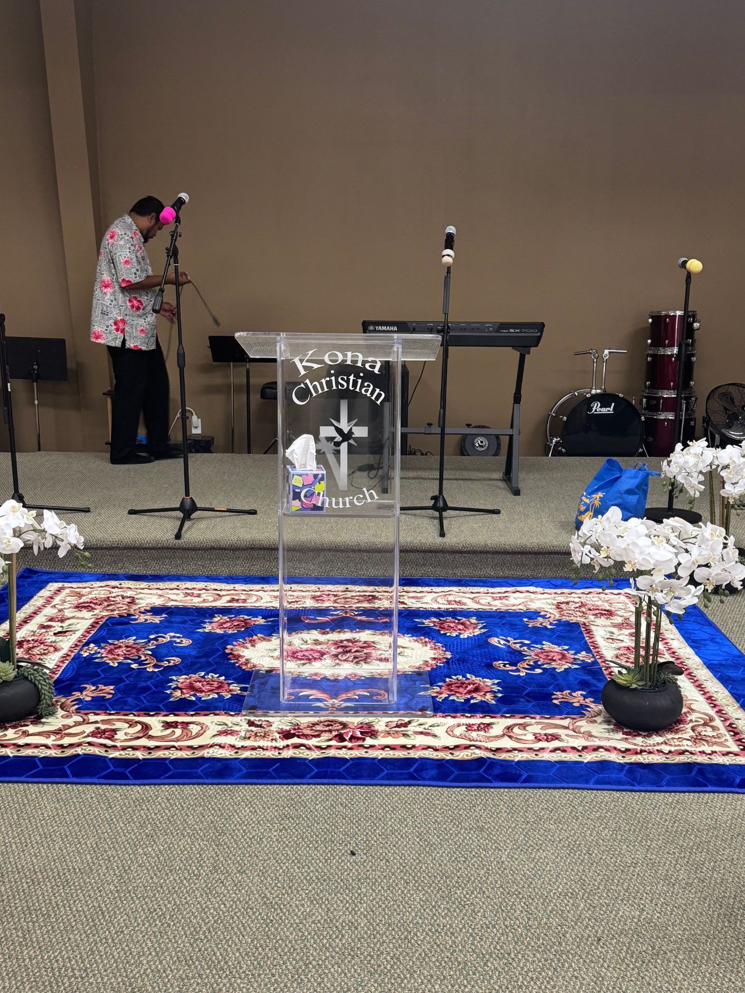 Stage with a podium, keyboard, drums, and a person setting up a microphone.