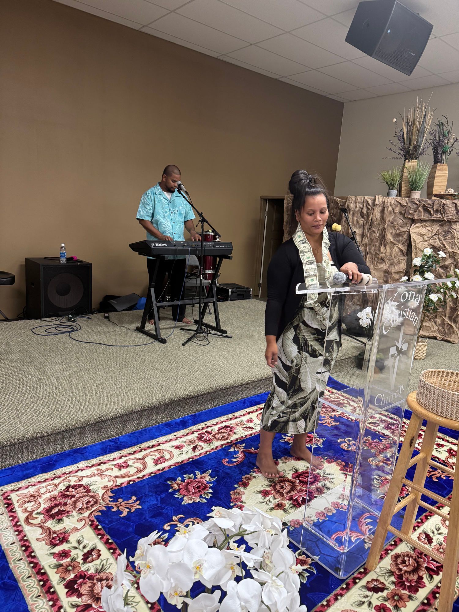 Woman at clear podium speaks; man plays keyboard on stage; floral carpet.