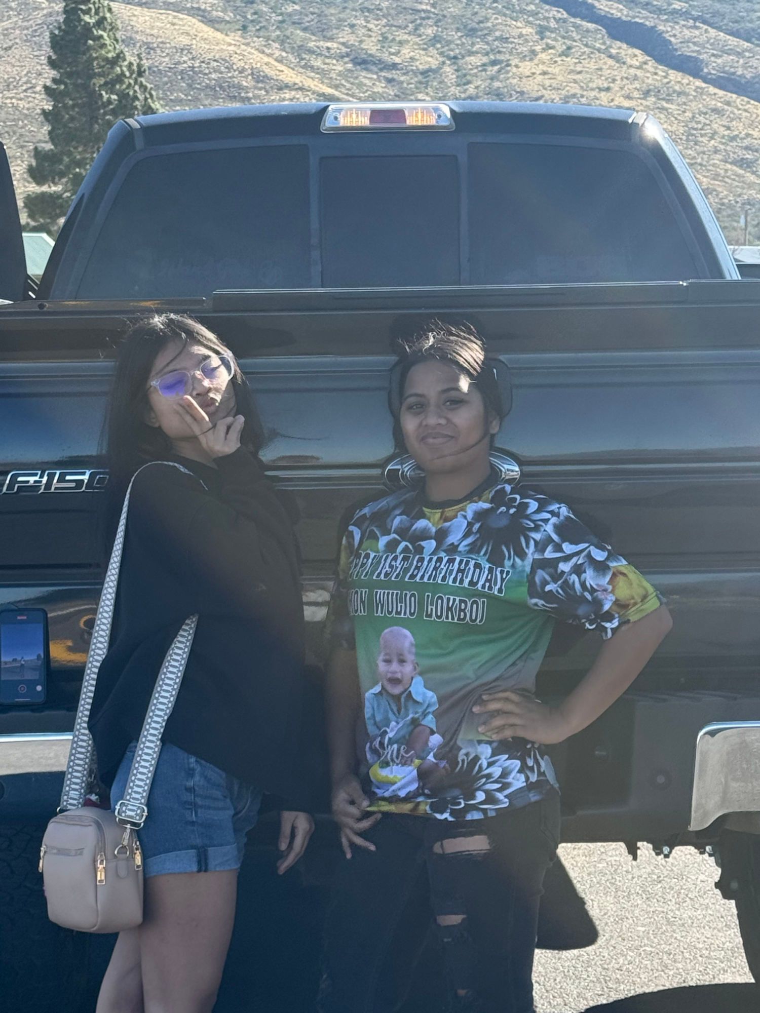 Two young women pose in front of a black pickup truck. One smiles, giving a peace sign, the other has her hand on her hip.