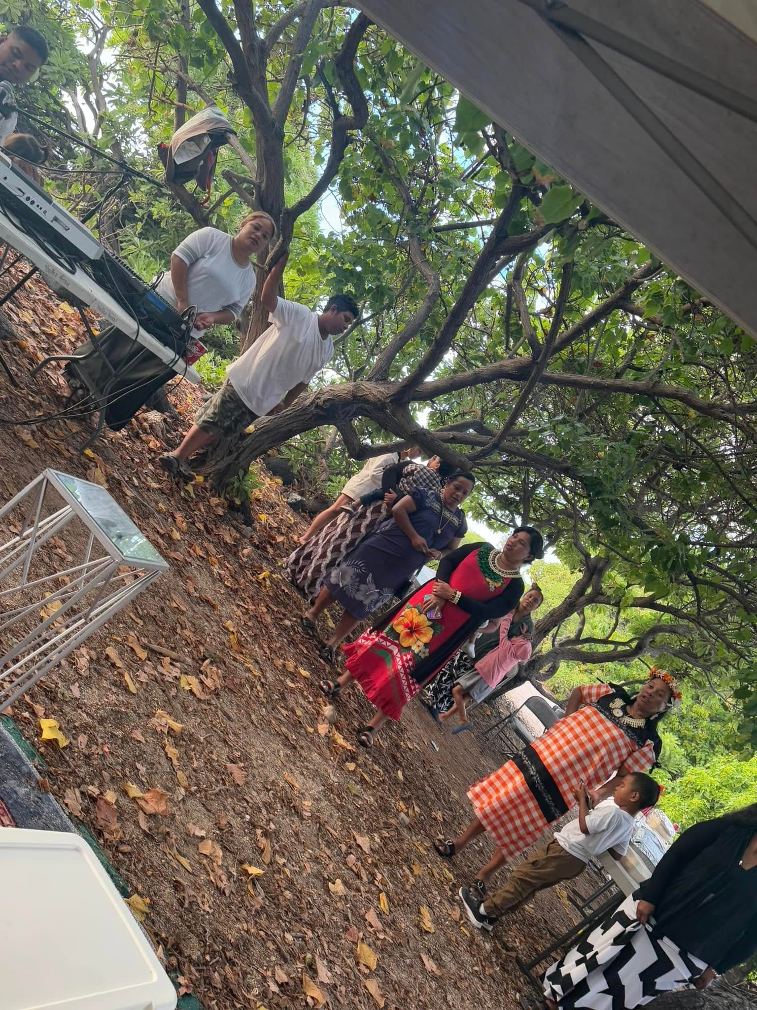 People under trees, wearing various clothes. Some standing, some sitting, enjoying an outdoor gathering.