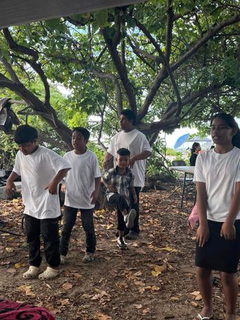 Group of people under a tree, some dancing. White shirts, dark pants. Outdoors.