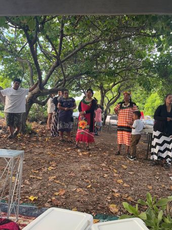 A group of people outdoors, some wearing patterned dresses, under trees; some are smiling.