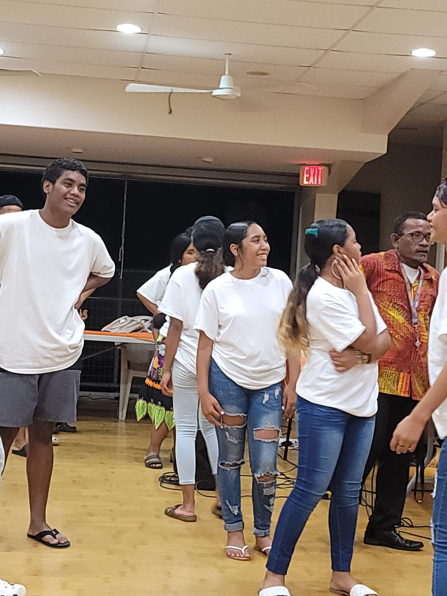 Group of people in white shirts indoors, smiling. Man in the back in a colorful shirt.