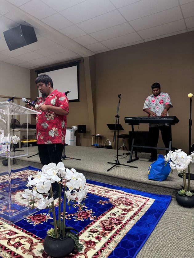 Two men in Hawaiian shirts on a church stage, one speaking at a podium, the other playing a keyboard.