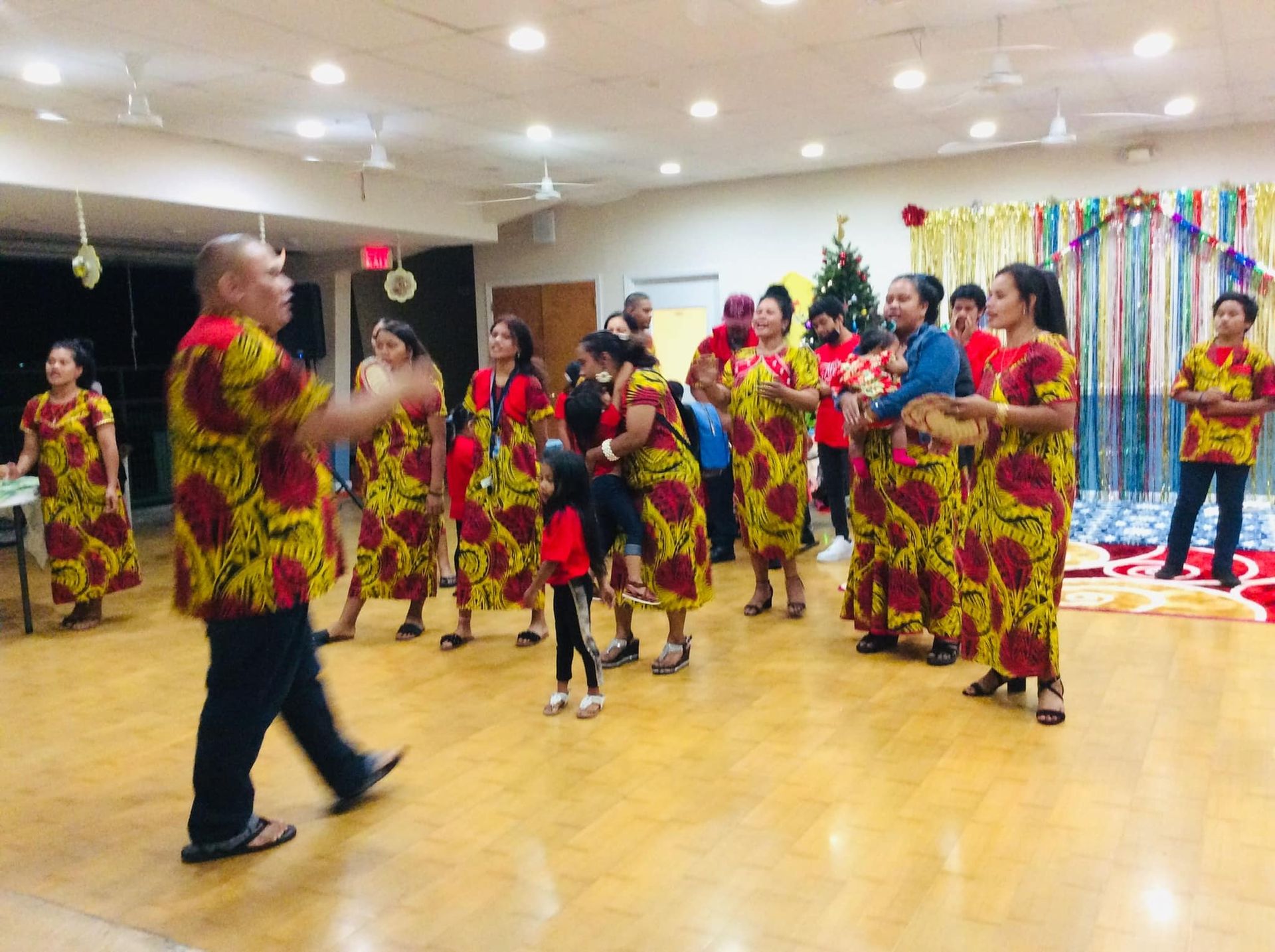 Group of people in patterned dresses dancing in a decorated hall; a man leads the dance.
