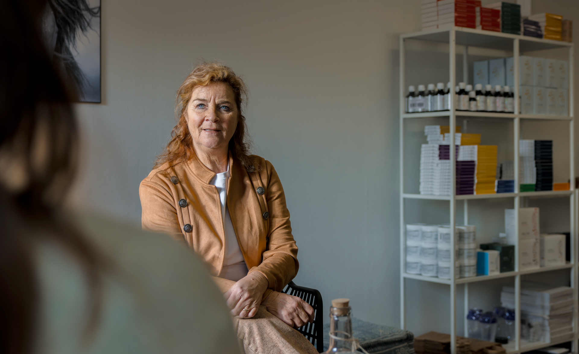 Een vrouw in een oranje vest zit tegenover een persoon en kijkt aandachtig. Achter haar staan ​​witte schappen met producten.