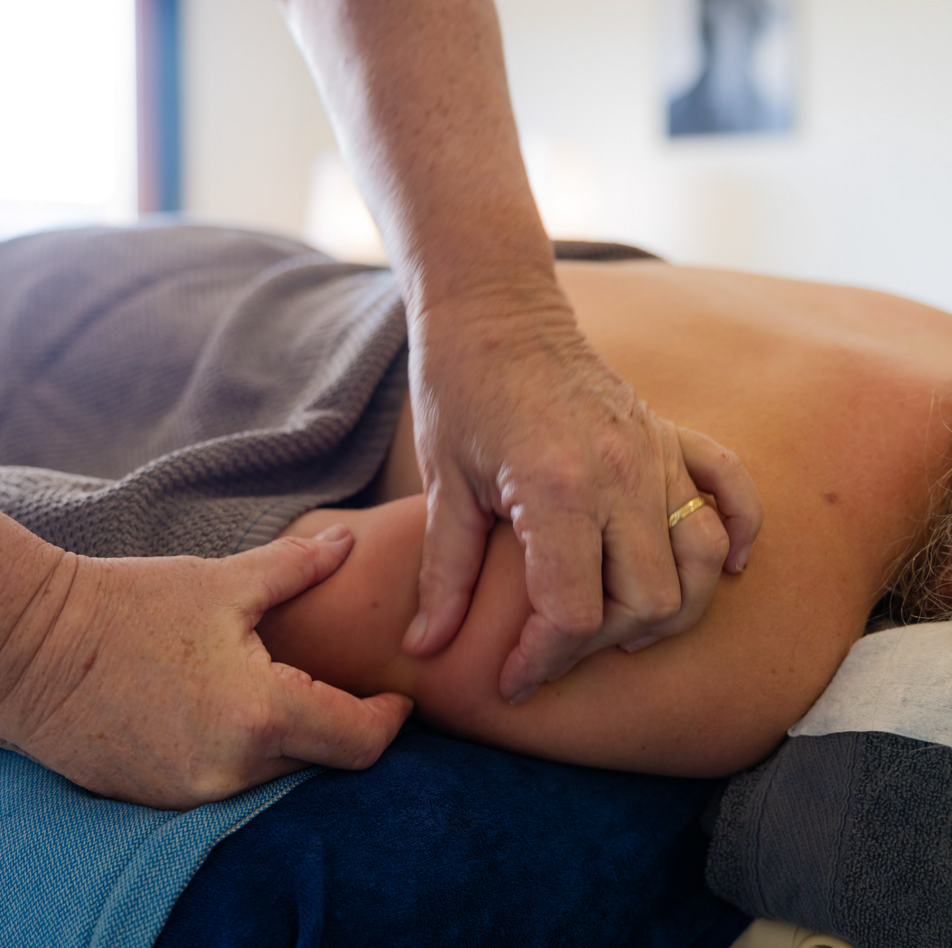 Hands massaging a person's upper arm, lying on a massage table, draped with a towel.