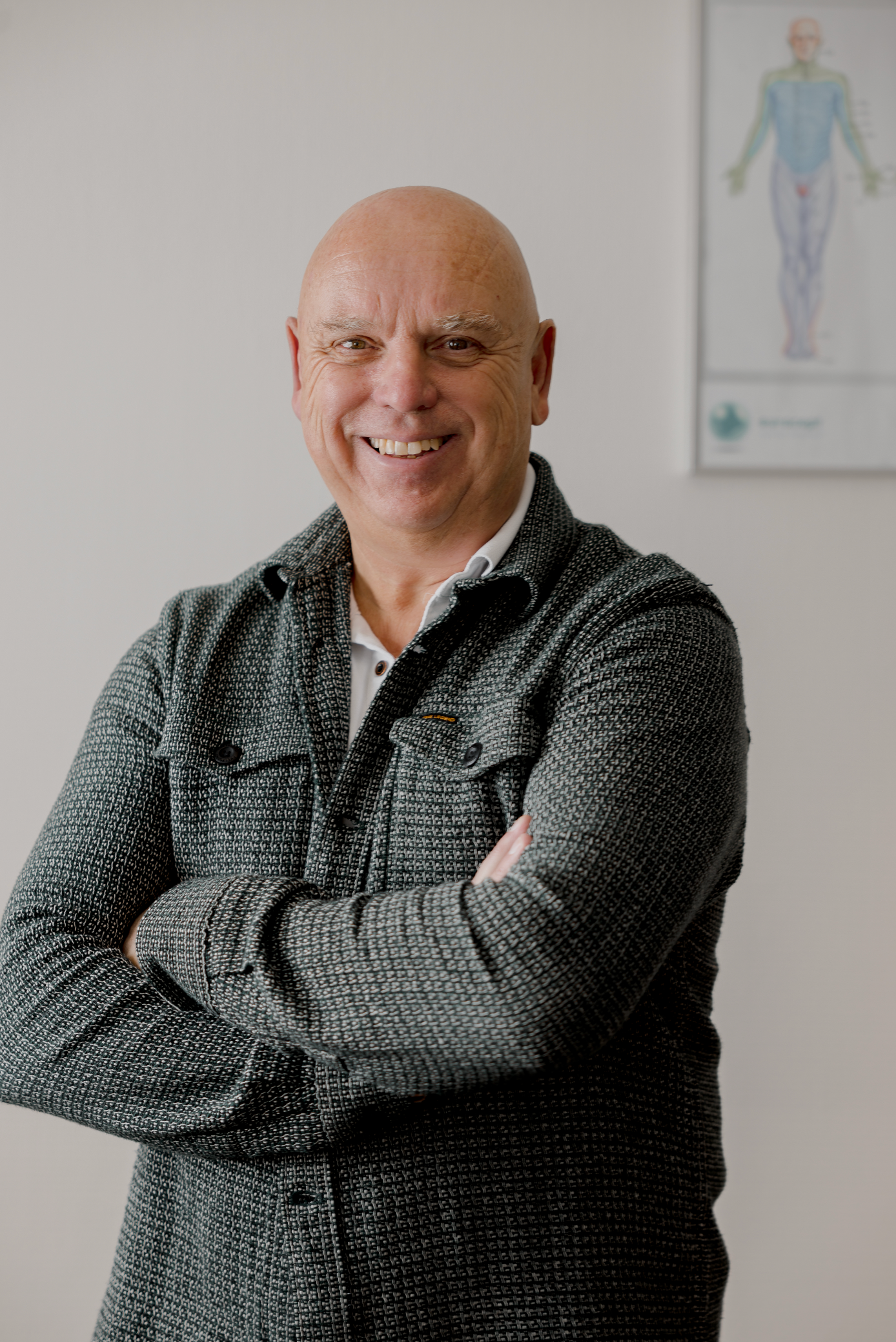 Een kale man met gekruiste armen, glimlachend, gekleed in een jas met patroon, staat in een kamer met een anatomische kaart.