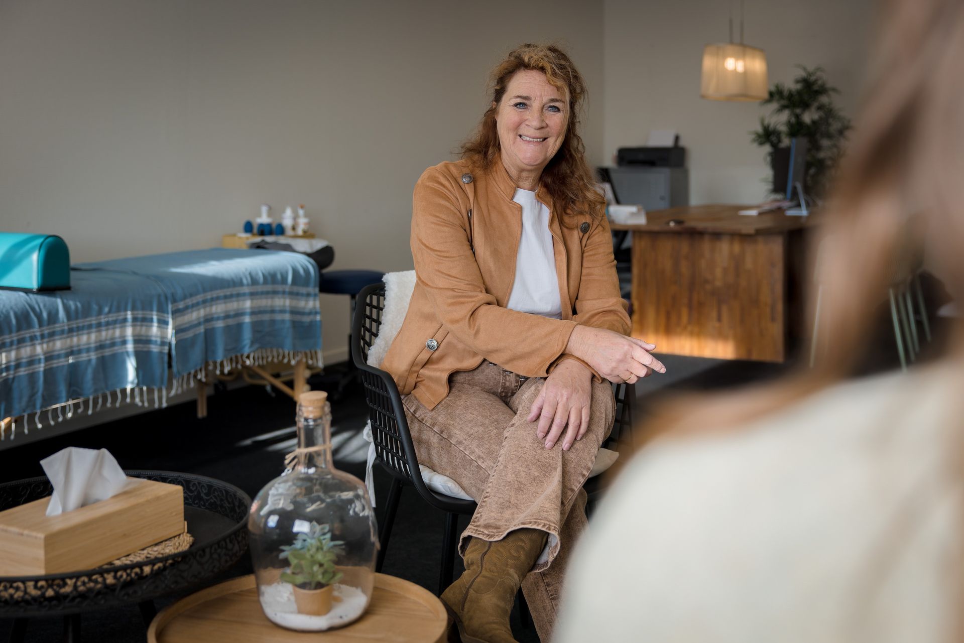 Een vrouw in een bruine jas glimlacht terwijl ze in een kantoor zit. 