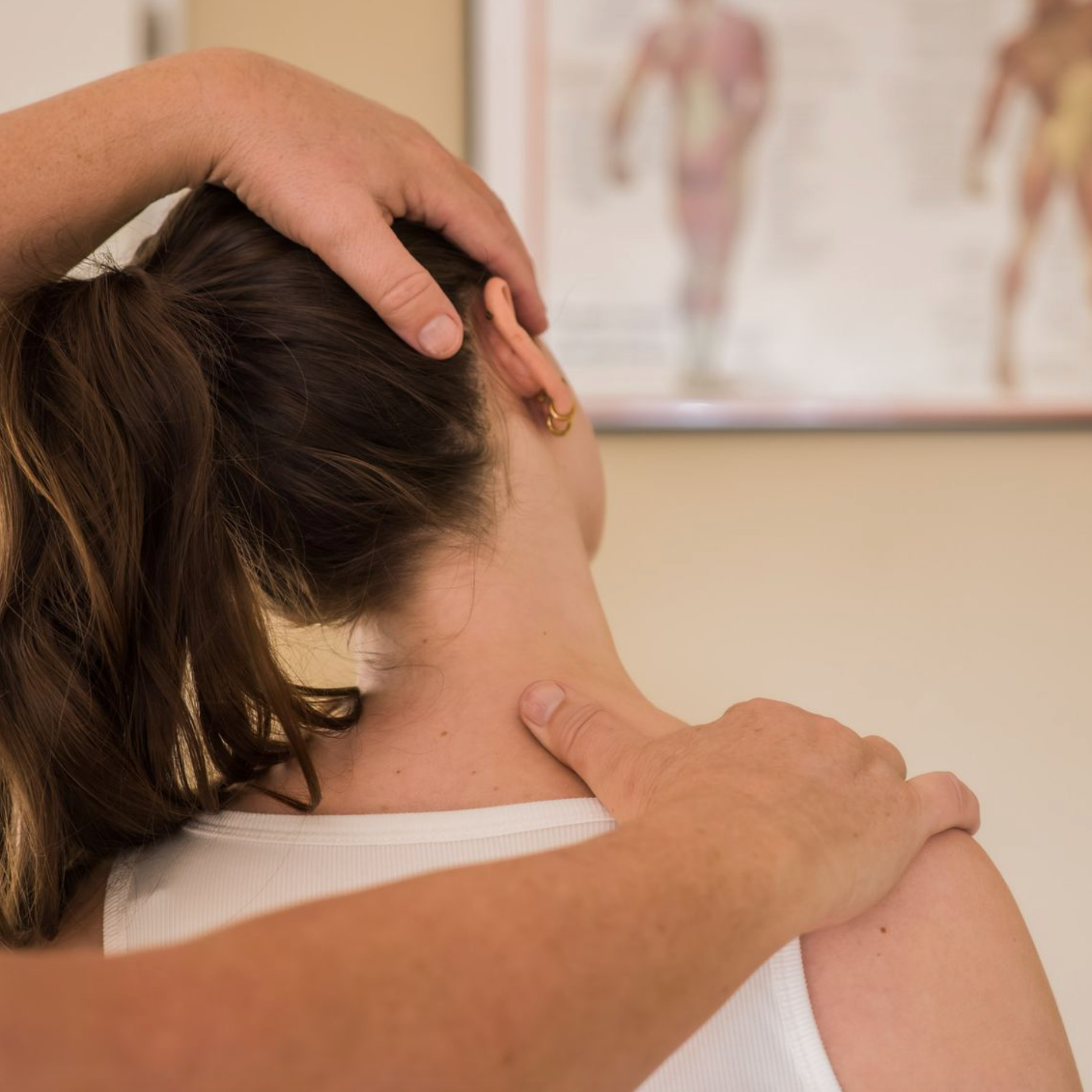 Handen die de nek en het hoofd van een persoon onderzoeken. Wit shirt, neutrale omgeving met een diagram aan de muur.