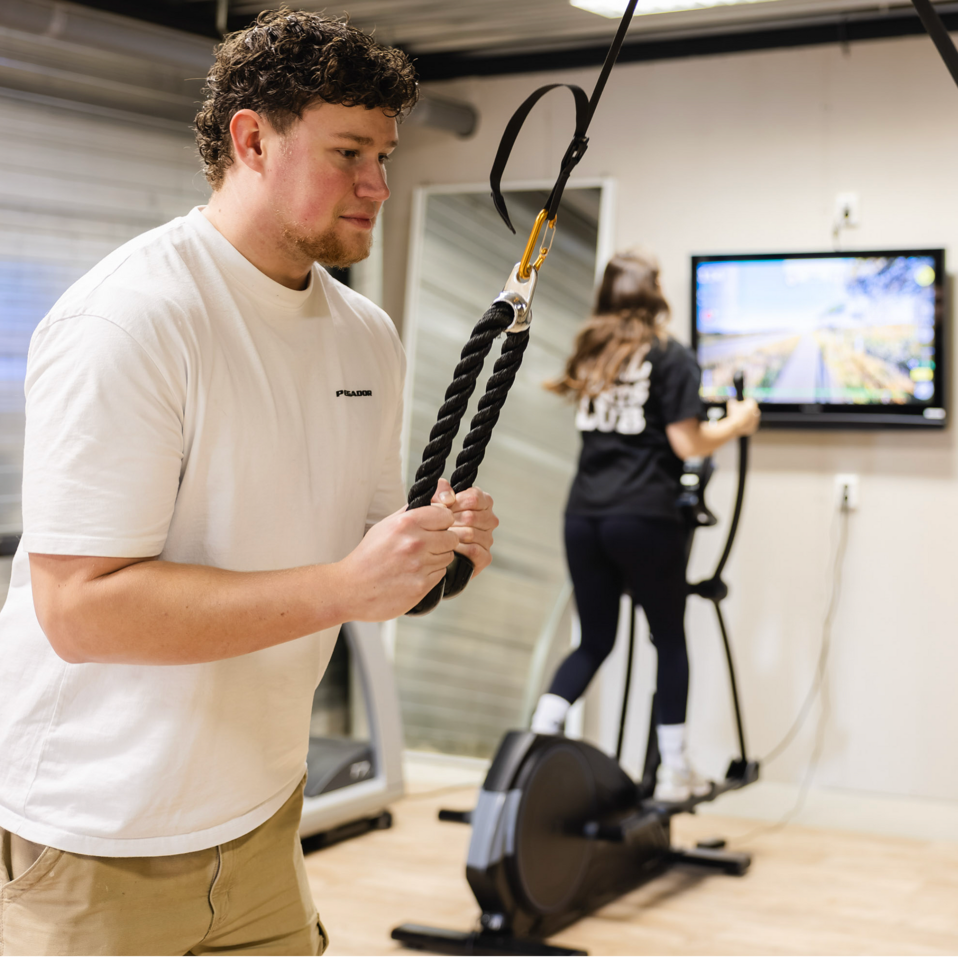 Man using rope machine, woman on elliptical in gym. White t-shirts, neutral-lit space, workout setting.