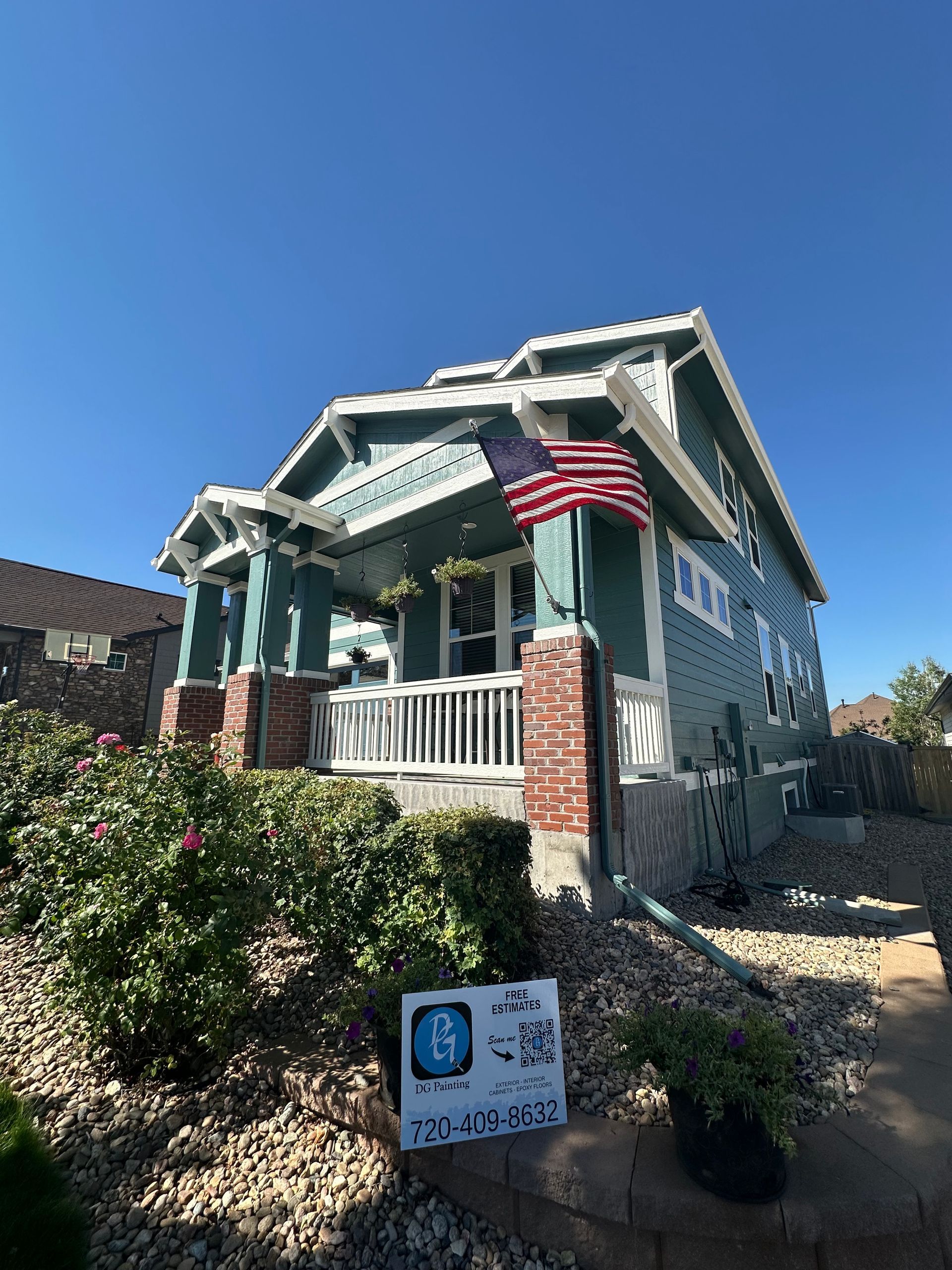 A teal house with brick pillars and a porch, featuring an American flag and a blue handicap-accessible sign in the yard.