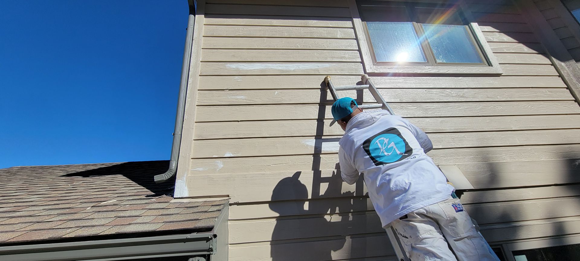 Person painting the side of a house while standing on a ladder in the sunlight.