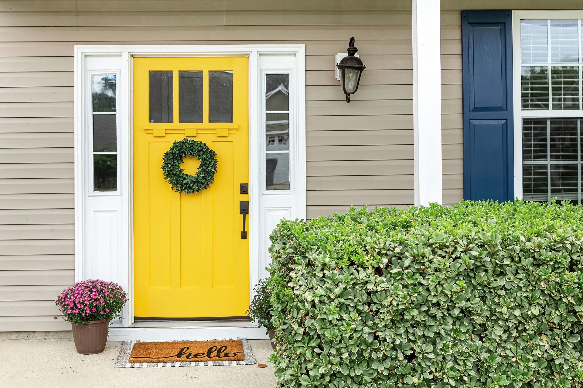 Yellow front door with a green wreath, flanked by white trim, with a potted plant and a 