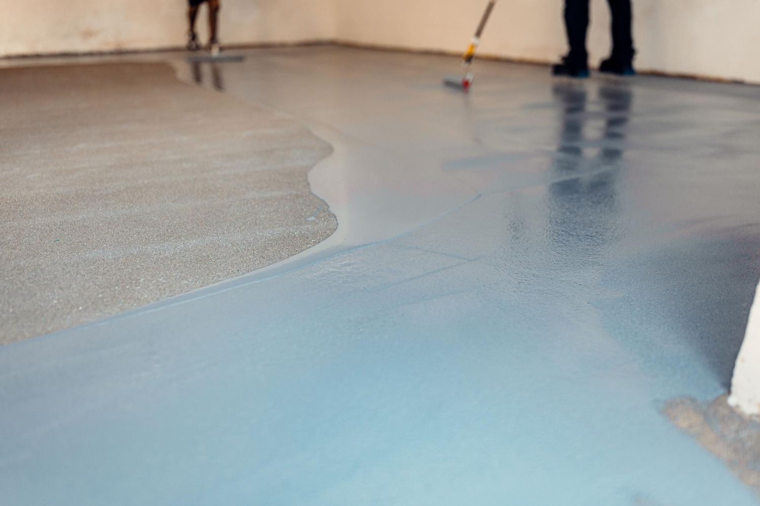Workers use rollers to apply a smooth, light blue epoxy coating over a gray concrete floor in an unfinished room.