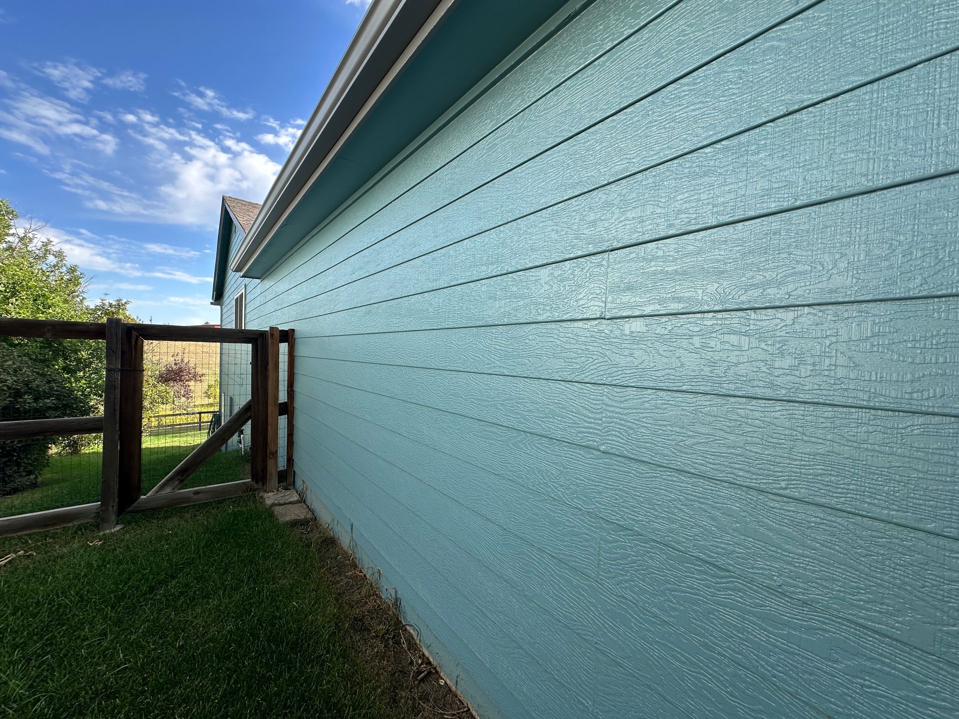 Blue house siding beside a grassy yard and wooden gate under a partly cloudy sky