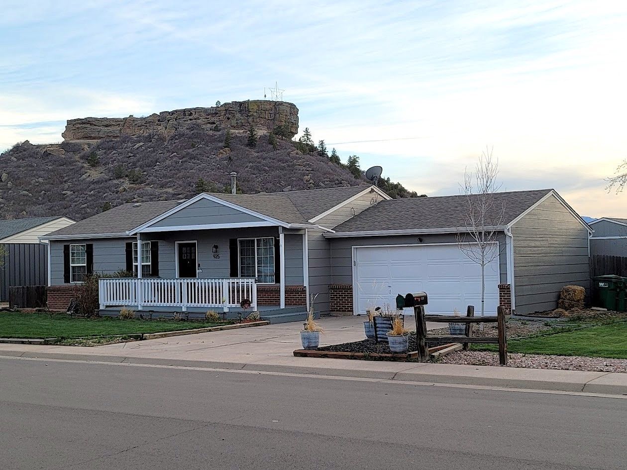 Gray house with a detached garage, set against a rocky hillside under a cloudy sky.