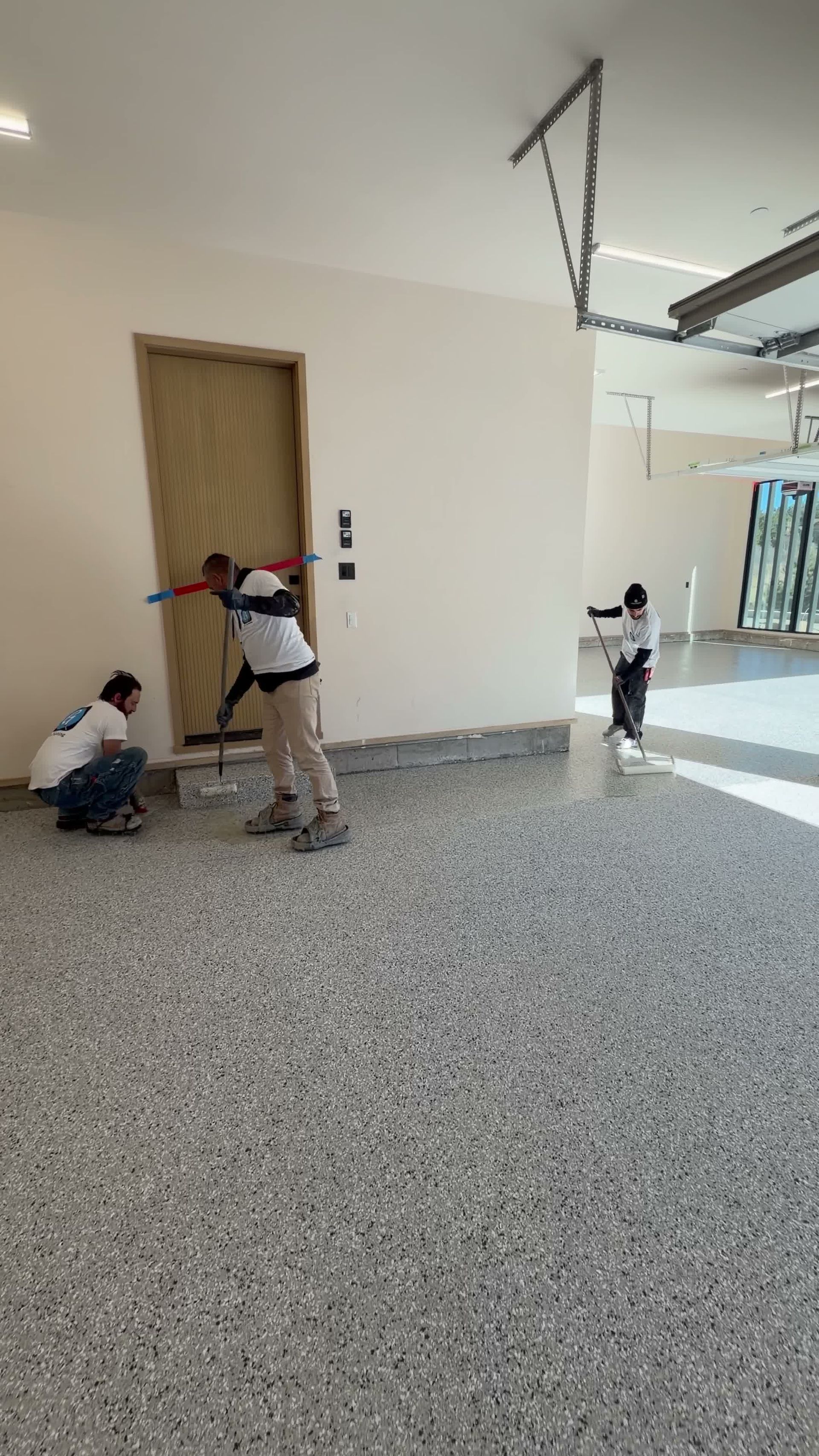 Three people apply epoxy flooring in a garage. One uses a roller on the floor, two work near a door.