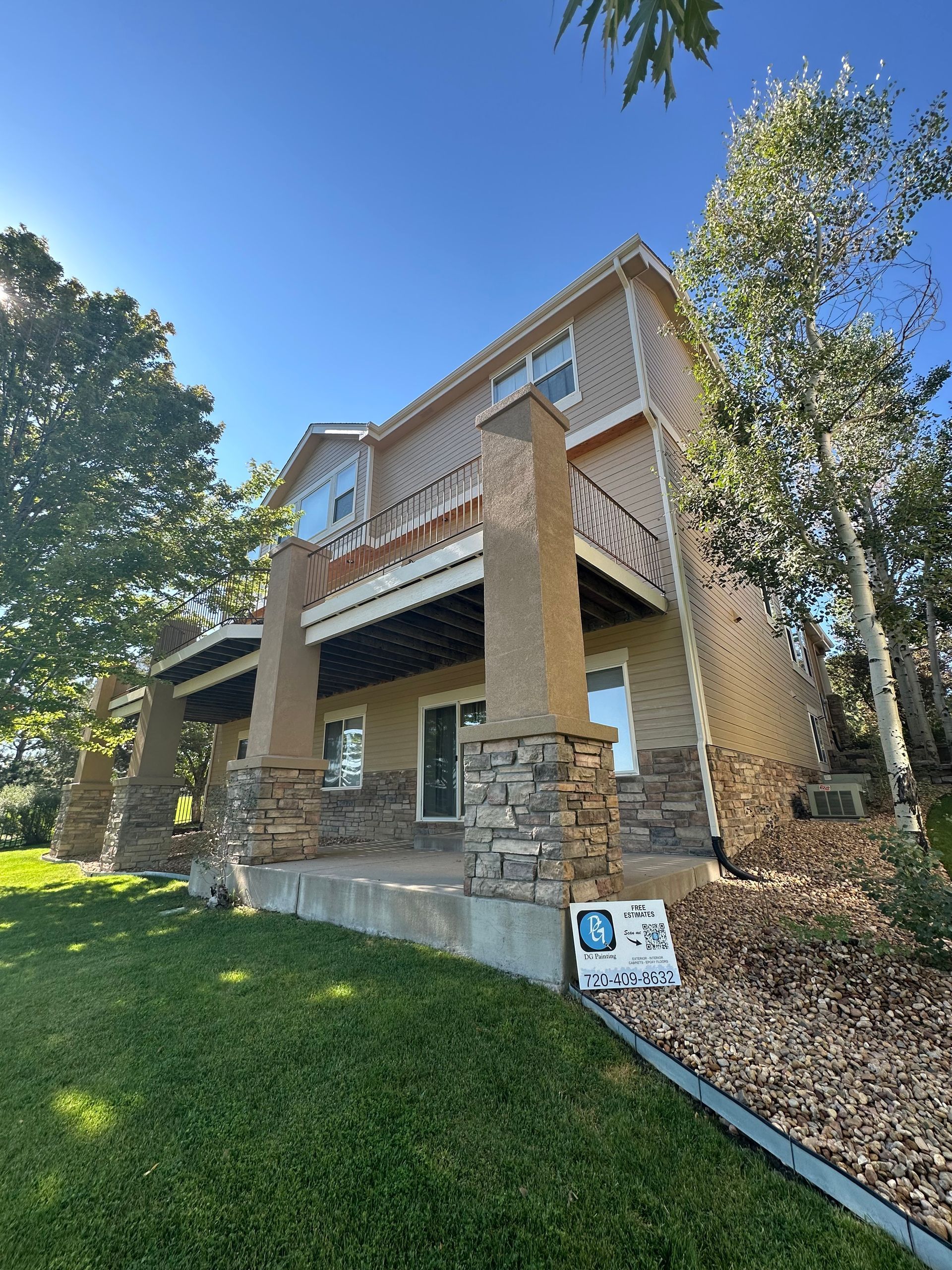 Two-story house with stone pillars, tan siding, and a wooden deck on a sunny day with grass and trees.
