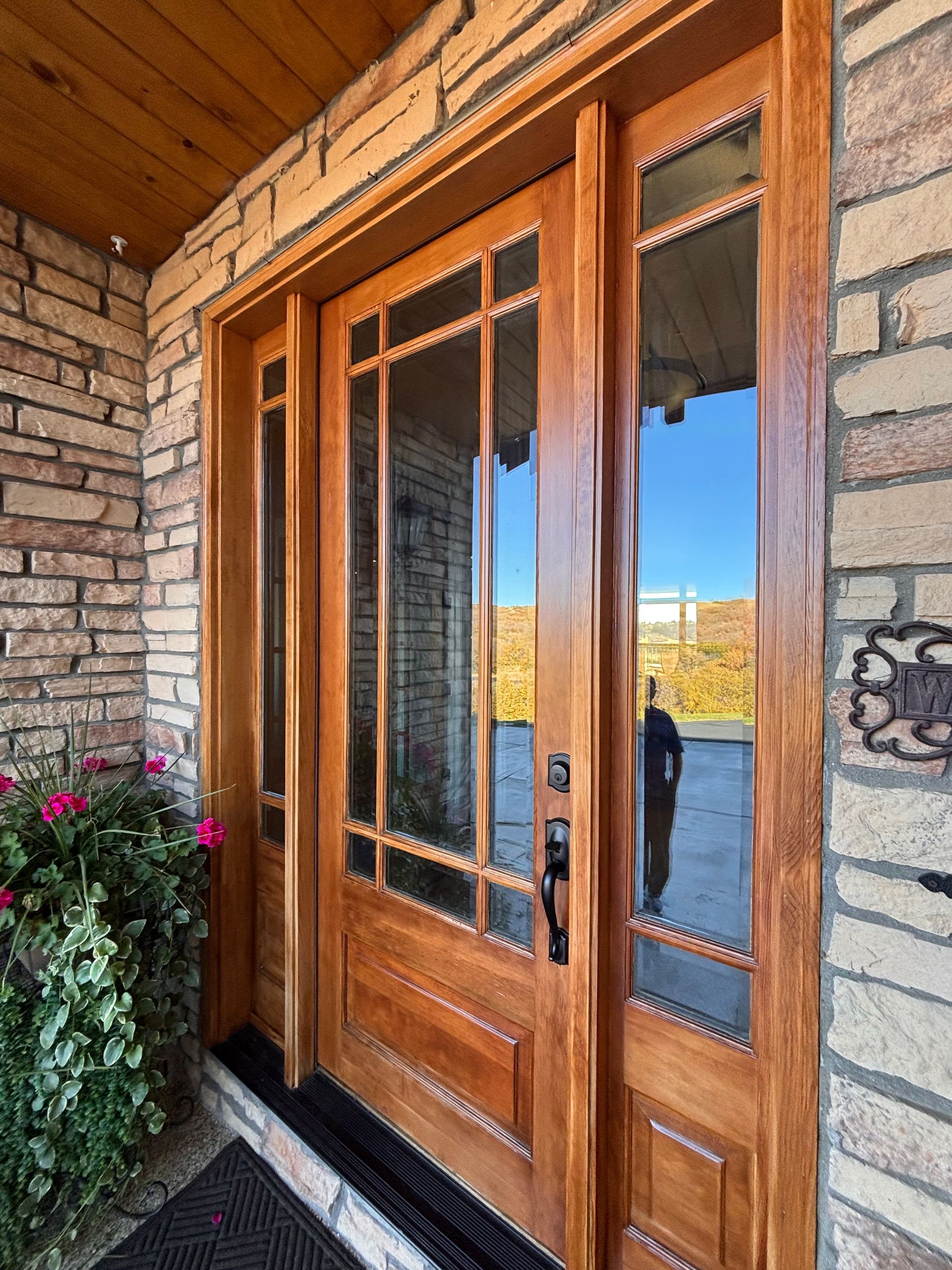 Wooden front door with glass panels; person reflected in side window, surrounded by stone facade.