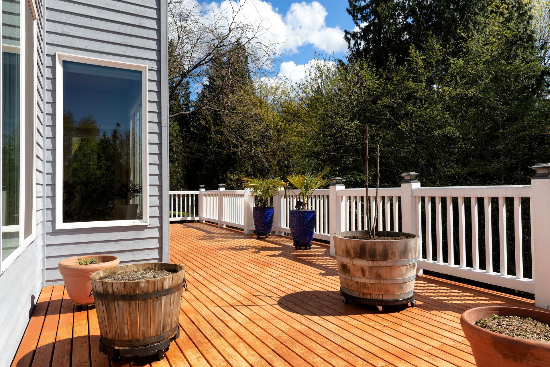 A sunlit wooden deck with potted plants and wooden barrel planters, framed by white railings and a grey house exterior.